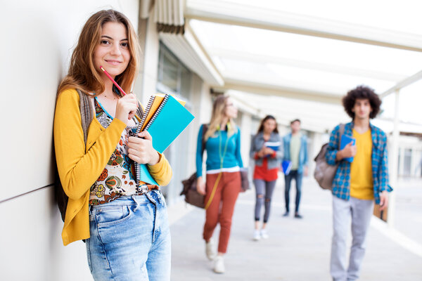 Eine junge Frau lehnt an einer Wand und h�lt Notizb�cher und Stift in den H�nden. Im Hintergrund laufen Jugendliche im Campus.