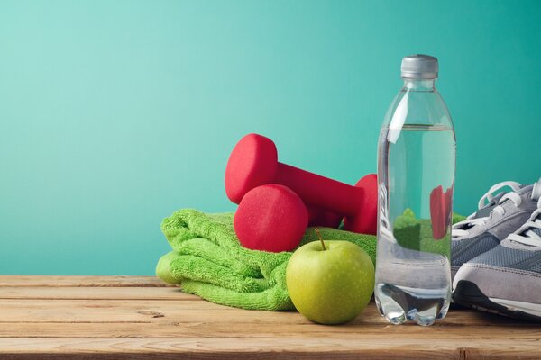 Wasserflasche, Apfel, Hanteln und Sportschuhe auf einem Holztisch vor t&uuml;rkisem Hintergrund