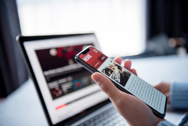 Eine Hand h�lt ein Smartphone, auf dem ein Online-Nachrichtendienst zu sehen ist. Im Hintergrund ist unscharf ein Laptop abgebildet.