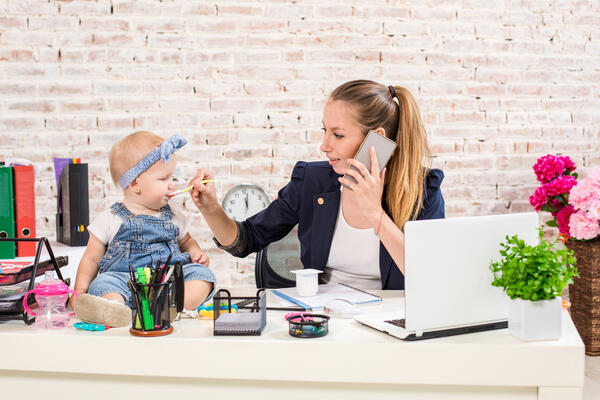 Geschäftsfrau und Mutter führt ein Telefongespräch am Arbeitsplatz und füttert dabei ihr kleines Kind.
