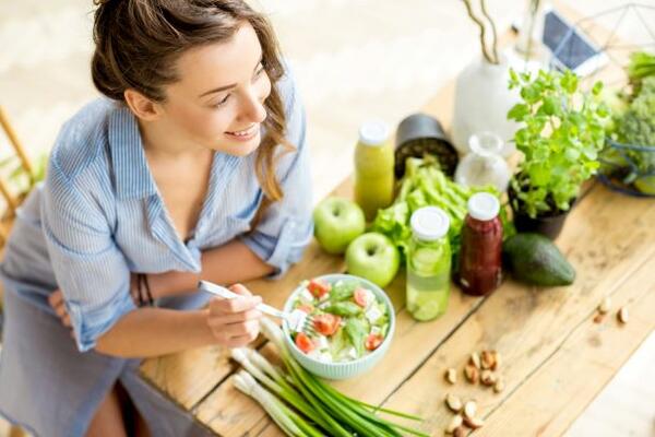 Junge und glückliche Frau sitzt am Tisch, auf dem frische grüne Zutaten stehen und isst einen gesunden Salat.